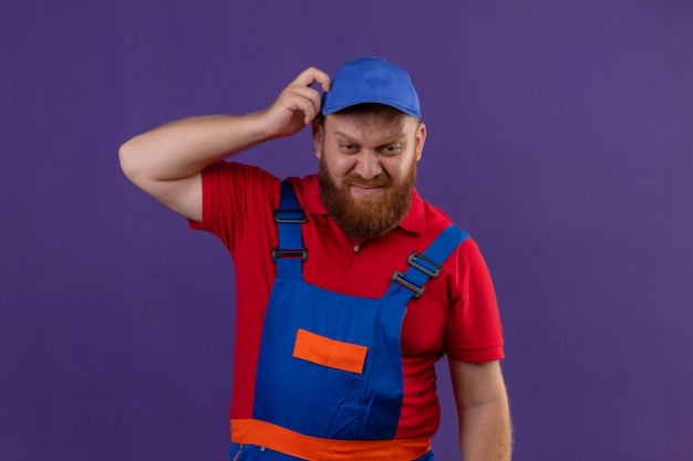 Hombre joven constructor barbudo en uniforme de construcción y gorra que parece incierto y confundido rascándose la cabeza sobre fondo púrpura