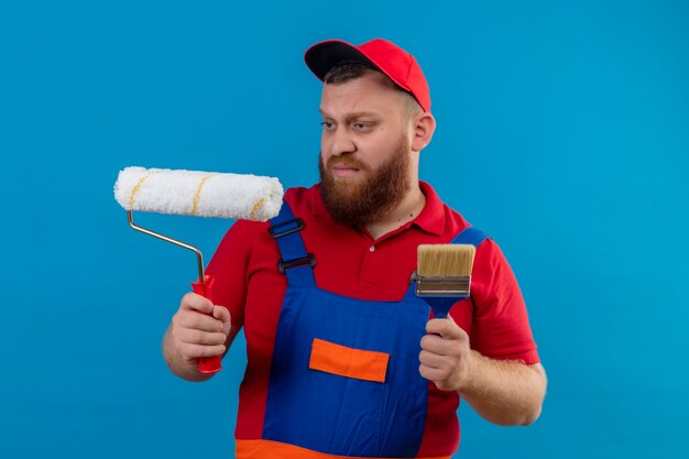 Hombre joven constructor barbudo en uniforme de construcción y gorra con rodillo de pintura y pincel mirando el rodillo con expresión escéptica en la cara