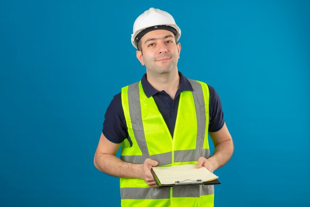 Hombre joven constructor con casco blanco y un chaleco amarillo, sosteniendo portapapeles en manos de pie en azul aislado