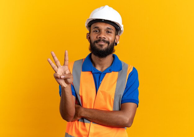 Hombre joven constructor complacido en uniforme con casco de seguridad gesticulando tres con los dedos aislado en la pared naranja con espacio de copia