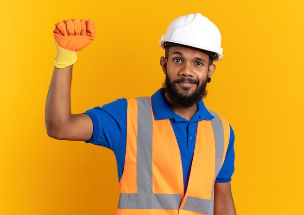 Hombre joven constructor complacido en uniforme con casco de seguridad y guantes de pie con el puño levantado aislado en la pared naranja con espacio de copia