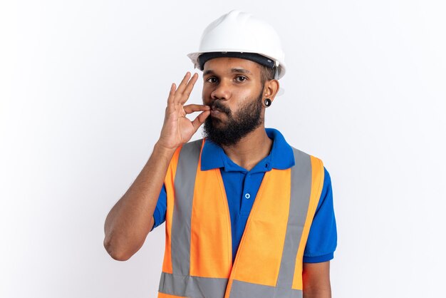 Hombre joven constructor complacido en uniforme con casco de seguridad haciendo un gesto perfecto aislado en la pared blanca con espacio de copia