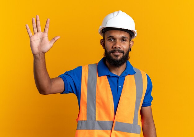 Hombre joven constructor complacido en uniforme con casco de seguridad de pie con la mano levantada aislado en la pared naranja con espacio de copia