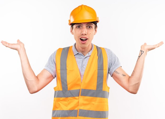 Hombre joven constructor complacido en uniforme extendiendo las manos aisladas en la pared blanca