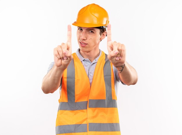 Hombre joven constructor complacido en uniforme mostrando uno aislado en la pared blanca