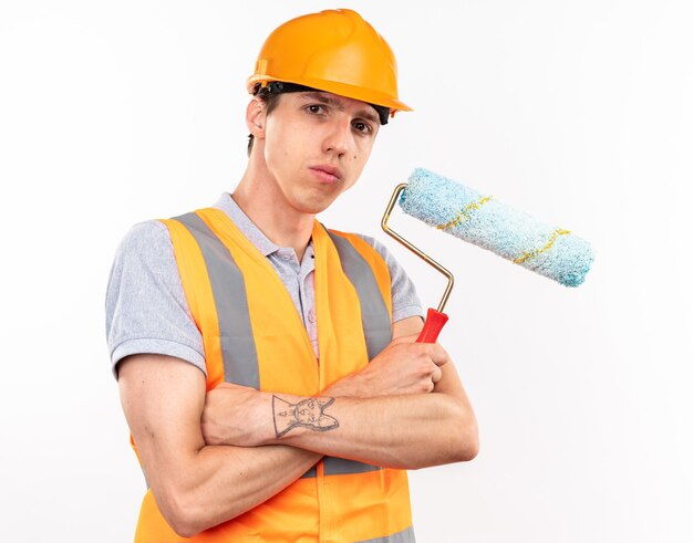 Hombre joven constructor confiado en uniforme cruzando las manos sosteniendo un cepillo de rodillo