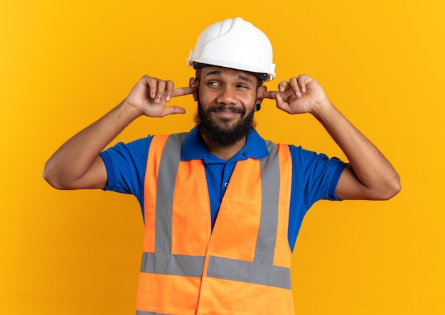 Foto gratuita hombre joven constructor descontento en uniforme con casco de seguridad cerrando sus oídos con los dedos mirando al lado aislado en la pared naranja con espacio de copia