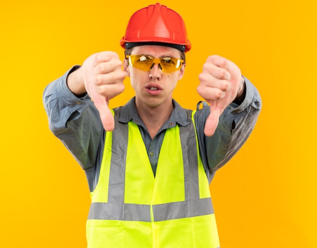 Hombre joven constructor disgustado en uniforme con gafas mostrando los pulgares hacia abajo aislado en la pared amarilla