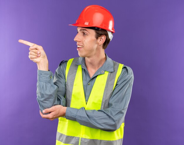 Hombre joven constructor de lado mirando complacido en puntos uniformes en el lado aislado en la pared azul con espacio de copia