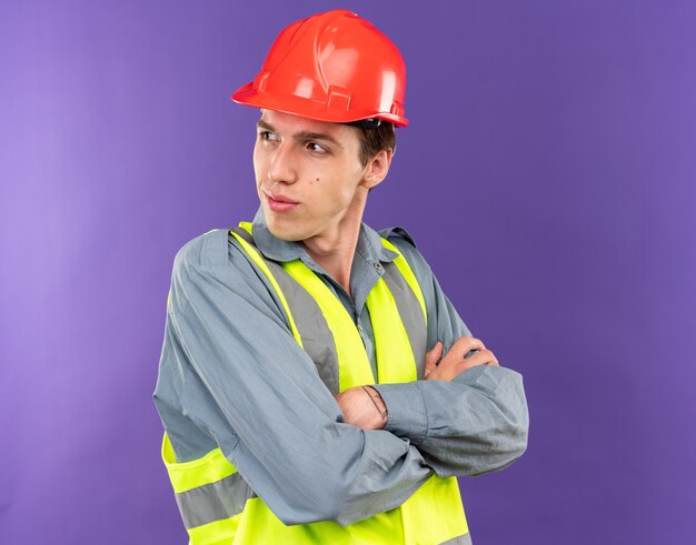 Hombre joven constructor de lado mirando complacido en uniforme cruzando las manos aisladas en la pared azul