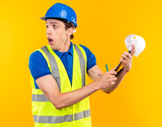 Foto gratuita hombre joven constructor de lado mirando preocupado en uniforme con portapapeles