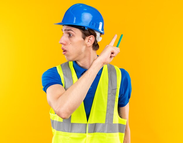 Hombre joven constructor de lado mirando sorprendido en uniforme sosteniendo lápiz y puntos detrás aislado en pared amarilla con espacio de copia