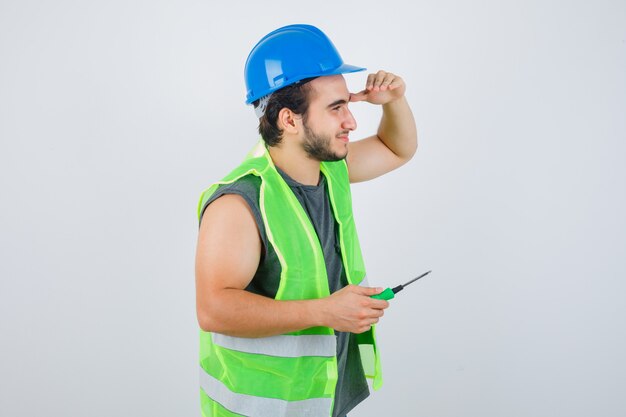 Hombre joven constructor manteniendo el destornillador mientras sostiene la mano en la cabeza para ver claramente en uniforme y lucir feliz. vista frontal.