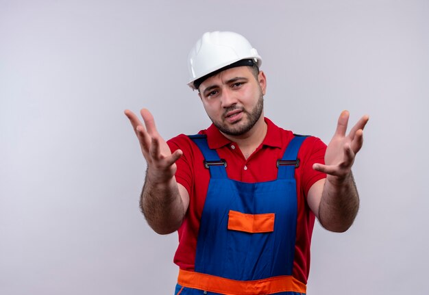 Hombre joven constructor molesto en uniforme de construcción y casco de seguridad sosteniendo las manos mirando a la cámara disgustado