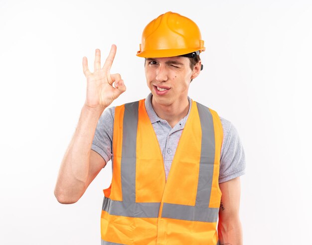 Hombre joven constructor parpadeó complacido en uniforme mostrando gesto bien aislado en la pared blanca