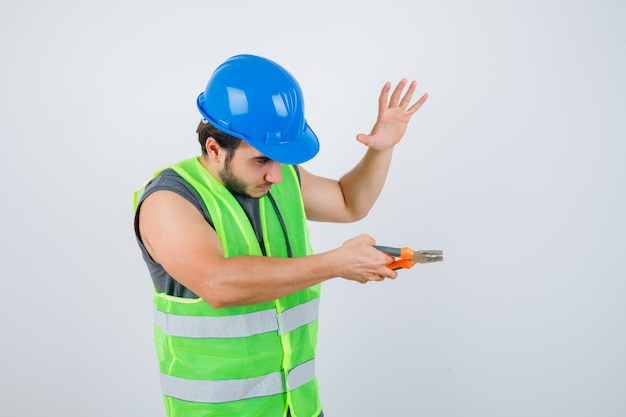 Hombre joven constructor sosteniendo alicates mientras levanta la palma en uniforme de ropa de trabajo y parece seguro de sí mismo. vista frontal.