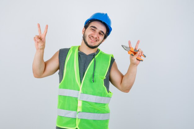 Hombre joven constructor sosteniendo alicates mientras muestra el signo de la victoria en uniforme de trabajo y luciendo alegre. vista frontal.