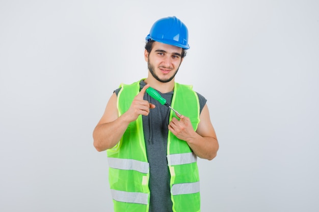 Hombre joven constructor sosteniendo un destornillador en uniforme y mirando confiado, vista frontal.