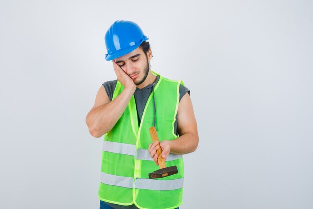 Hombre joven constructor sosteniendo un martillo mientras se inclina la mejilla en la mano en uniforme de trabajo y mirando pensativo, vista frontal.