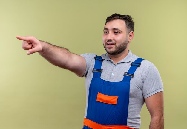 Hombre joven constructor en uniforme de construcción apuntando con el dedo índice a algo sonriendo