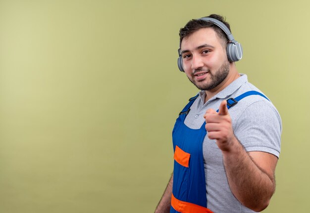 Hombre joven constructor en uniforme de construcción con auriculares sonriendo con cara feliz apuntando con el dedo índice a la cámara