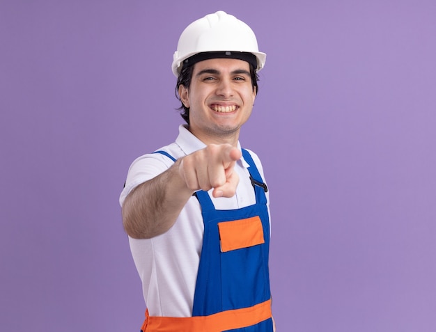 Hombre joven constructor en uniforme de construcción y casco de seguridad apuntando con el dedo índice al frente sonriendo alegremente de pie sobre la pared púrpura