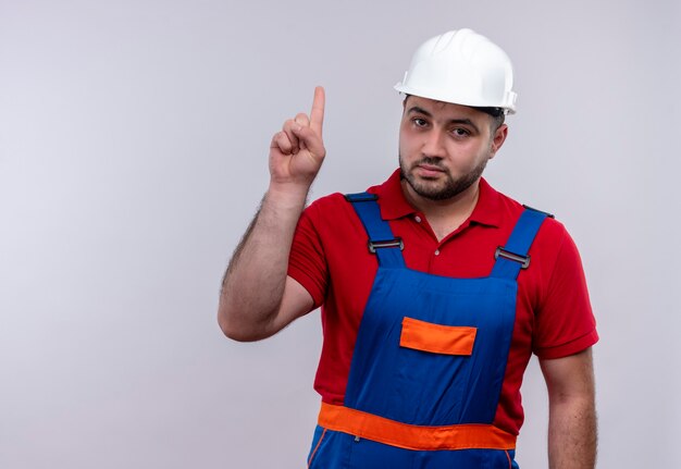 Hombre joven constructor en uniforme de construcción y casco de seguridad apuntando con el dedo índice hacia arriba mirando confiado