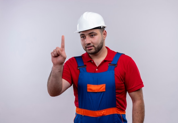 Hombre joven constructor en uniforme de construcción y casco de seguridad apuntando con el dedo índice hacia arriba mirando confiado