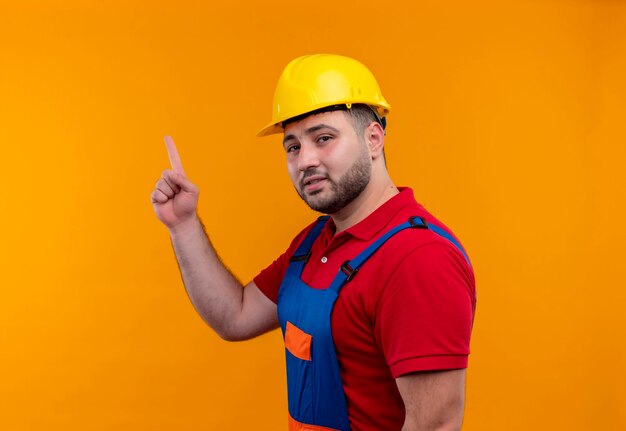 Hombre joven constructor en uniforme de construcción y casco de seguridad apuntando con el dedo índice hacia arriba mirando inteligente y seguro centrado en la tarea