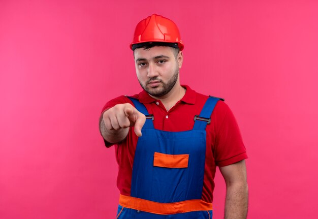 Hombre joven constructor en uniforme de construcción y casco de seguridad apuntando con índice a la cámara disgustado