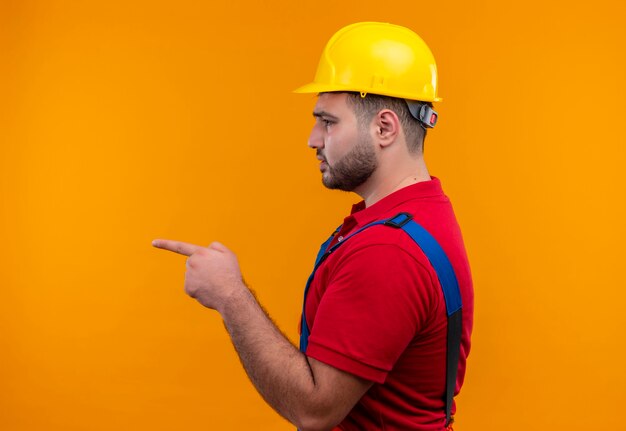 Hombre joven constructor en uniforme de construcción y casco de seguridad apuntando hacia los lados con el dedo índice a algo o alguien con el ceño fruncido serio