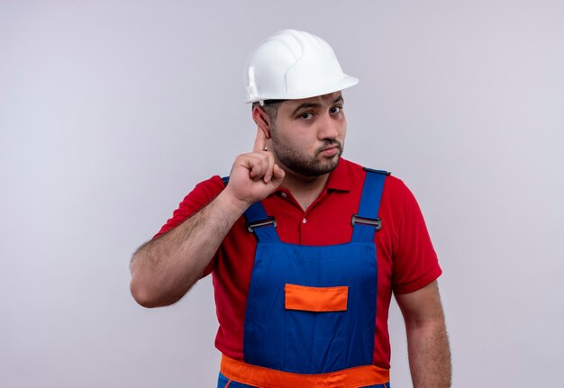 Hombre joven constructor en uniforme de construcción y casco de seguridad apuntando a su oído tratando de escuchar