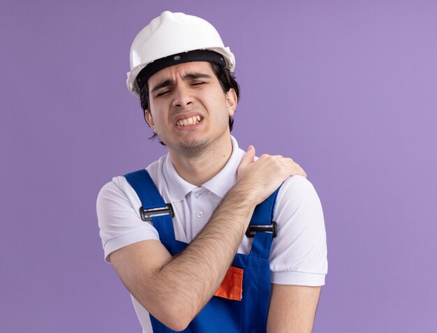 Hombre joven constructor en uniforme de construcción y casco de seguridad con aspecto de malestar tocando su hombro sintiendo dolor de pie sobre la pared púrpura