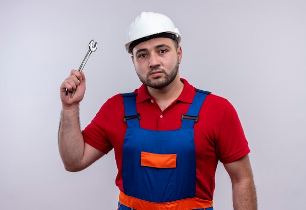 Foto gratuita hombre joven constructor en uniforme de construcción y casco de seguridad balanceando un martillo con cara seria
