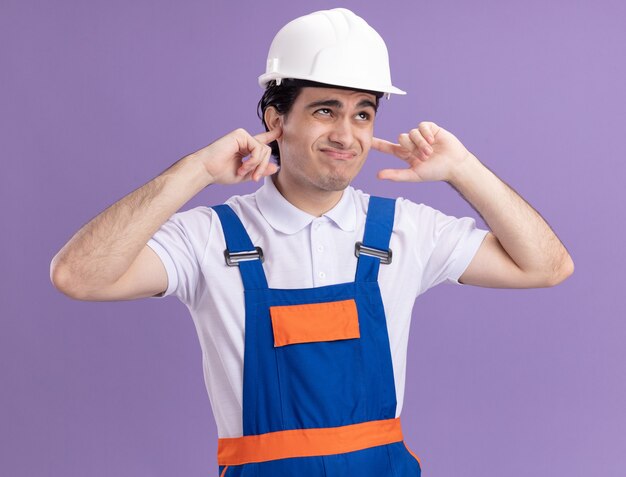 Hombre joven constructor en uniforme de construcción y casco de seguridad cerrando sus oídos con los dedos con expresión molesta de pie sobre la pared púrpura