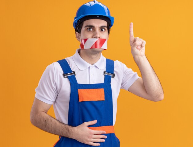 Hombre joven constructor en uniforme de construcción y casco de seguridad con cinta envuelta alrededor de la boca mirando al frente preocupado apuntando con el dedo índice sobre la pared naranja