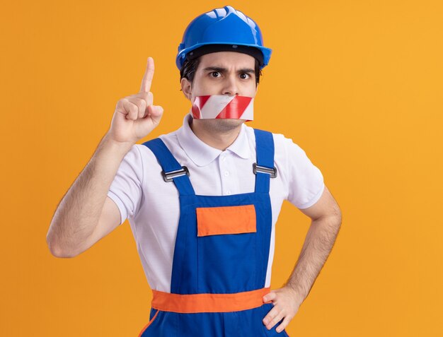 Hombre joven constructor en uniforme de construcción y casco de seguridad con cinta envuelta alrededor de la boca mirando al frente preocupado apuntando con el dedo índice sobre la pared naranja