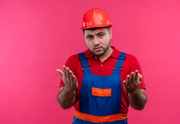 Hombre joven constructor en uniforme de construcción y casco de seguridad disgustado argumentando gesticulando con las manos