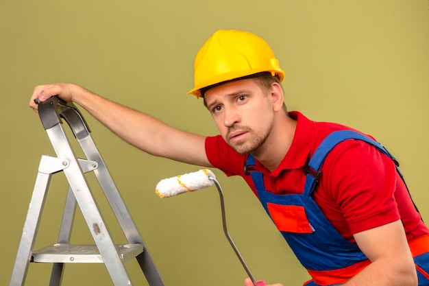 Hombre joven constructor en uniforme de construcción y casco de seguridad en escalera de metal con rodillo de pintura mirando hacia los lados sobre la pared verde aislada