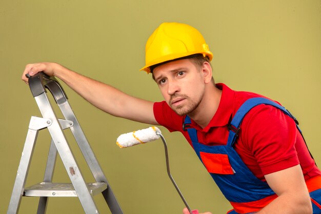 Hombre joven constructor en uniforme de construcción y casco de seguridad en escalera de metal con rodillo de pintura mirando hacia los lados sobre la pared verde aislada