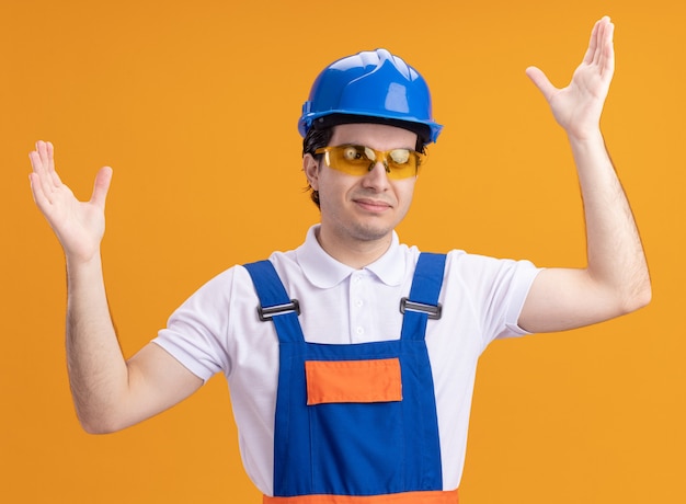 Hombre joven constructor en uniforme de construcción y casco de seguridad con gafas mirando al frente feliz y positivo con los brazos levantados de pie sobre la pared naranja