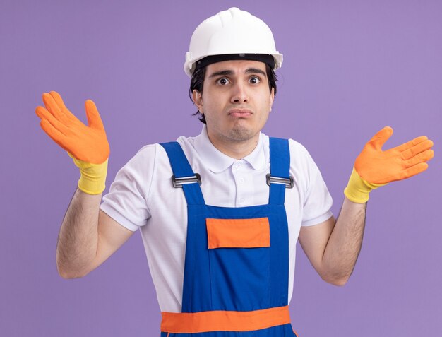 Hombre joven constructor en uniforme de construcción y casco de seguridad con guantes de goma mirando al frente confundido e inseguro con los brazos levantados sobre la pared púrpura