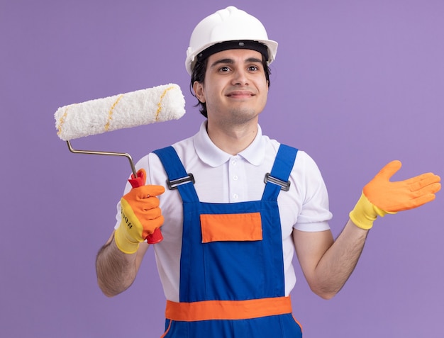 Hombre joven constructor en uniforme de construcción y casco de seguridad en guantes de goma con rodillo de pintura mirando a un lado con una sonrisa en la cara de pie sobre la pared púrpura