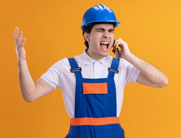 Hombre joven constructor en uniforme de construcción y casco de seguridad hablando por teléfono móvil gritando con expresión agresiva enojado y loco loco parado sobre pared naranja