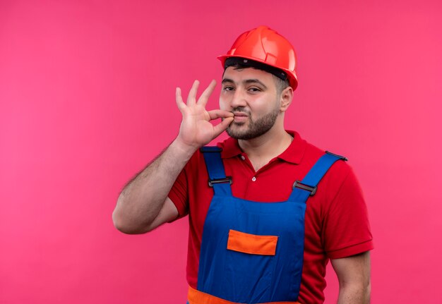 Hombre joven constructor en uniforme de construcción y casco de seguridad haciendo gesto de silencio como cerrar la boca con cremallera