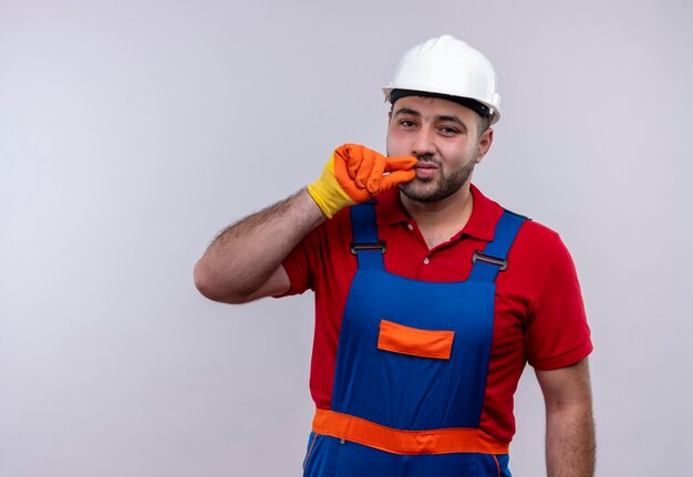 Hombre joven constructor en uniforme de construcción y casco de seguridad haciendo gesto de silencio como cerrar la boca con cremallera