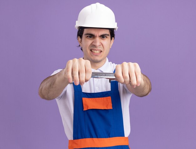 Hombre joven constructor en uniforme de construcción y casco de seguridad con llave mirándolo con cara enojada de pie sobre la pared púrpura