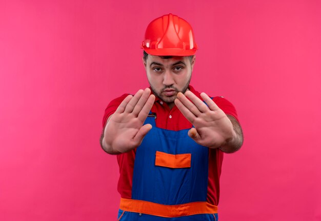 Hombre joven constructor en uniforme de construcción y casco de seguridad con las manos abiertas haciendo señal de stop con cara seria