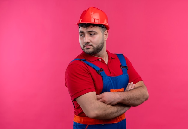 Hombre joven constructor en uniforme de construcción y casco de seguridad con las manos en el pecho cruzadas mirando a un lado con cara seria