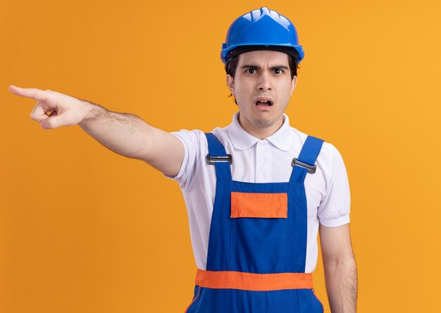 Hombre joven constructor en uniforme de construcción y casco de seguridad mirando al frente confundido y preocupado apuntando con el dedo índice a algo parado sobre la pared naranja